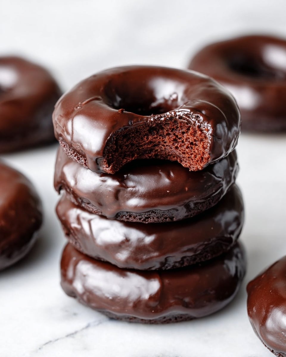 The image shows a close-up of a chocolate donut with a glossy, smooth dark chocolate glaze covering the entire top layer, dripping slightly down the sides. The donut is thick and rich, with a soft, moist, dark brown cake layer visible where a bite has been taken out on one side. The background is a white marbled texture that makes the dark chocolate donut stand out. Another similar donut slightly blurred sits in the foreground and background, creating depth. Photo taken with an iphone --ar 4:5 --v 7