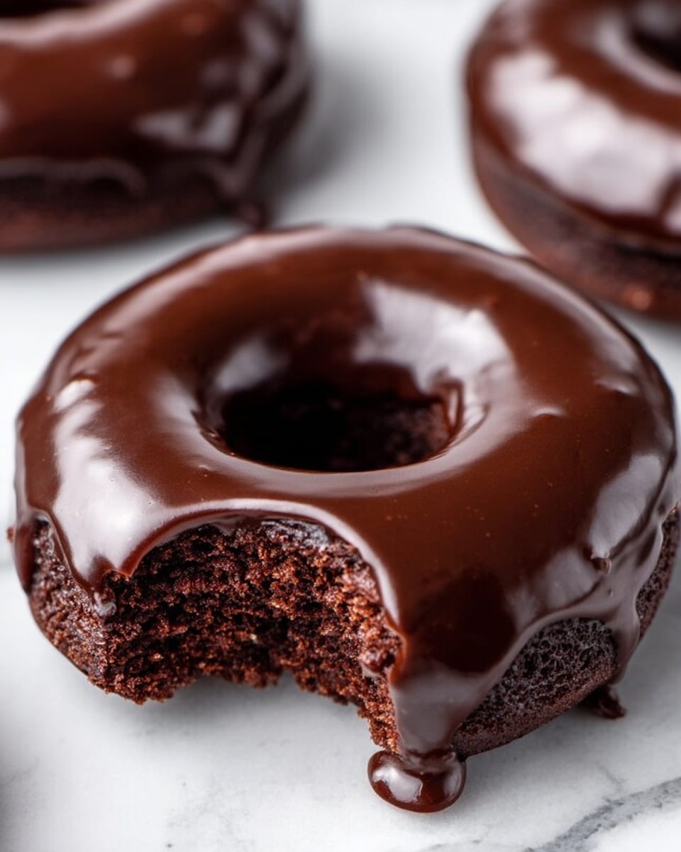 A stack of three chocolate donuts sits on a white marbled surface, each donut covered in a thick, shiny dark chocolate glaze. The top donut has a bite taken out of it, showing a soft, moist chocolate cake inside with a rich brown color. Around the stack, there are three more similarly glazed chocolate donuts blurred in the background, emphasizing the focus on the stack. The glaze is smooth, slightly cracked, and glossy, with a dark, almost black tone. photo taken with an iphone --ar 4:5 --v 7