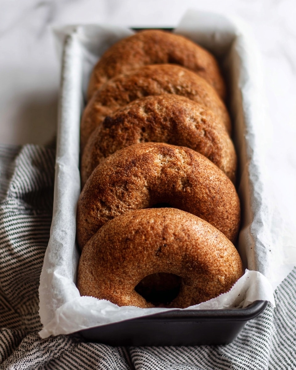 The image shows four brown bagels with a rough and slightly cracked texture, placed vertically in a black rectangular loaf pan lined with white parchment paper. The bagels have a dense, chewy appearance with a golden-brown crust, arranged one behind the other, filling the pan. The loaf pan is resting on a white marbled surface covered partially with a white cloth patterned with dark gray stripes. The focus is on the nearest bagel in the front with the others gradually fading into the background, showing a cozy and fresh baked look. photo taken with an iphone --ar 4:5 --v 7