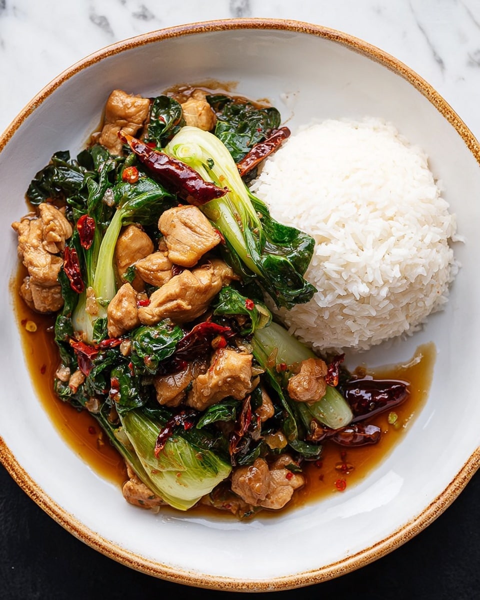 A white bowl filled with two layers of food, the right half holding a mound of plain white rice with a fluffy texture. On the left half, a stir-fry of browned chicken pieces mixed with bright green bok choy leaves and stems, dark green spinach or basil leaves, and deep red dried chili peppers in a shiny, brown sauce that pools slightly at the bottom. The edges of the bowl are slightly speckled with a natural, worn brown rim. The bowl is set on a white marbled surface. Photo taken with an iphone --ar 4:5 --v 7