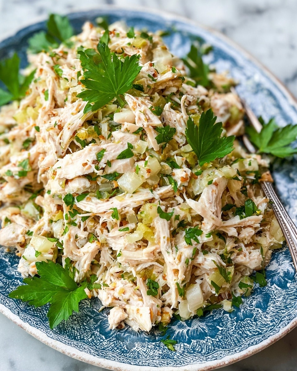 A white patterned oval plate holds a shredded chicken salad mixed with small pieces of light green celery, pale yellow artichoke hearts, and dark green herbs, all speckled with small red bits that look like dried chili flakes; the texture is moist but slightly chunky, with bright fresh parsley leaves scattered on top for color, and a vintage silver fork rests on the left side of the plate. The plate sits on a white marbled surface. photo taken with an iphone --ar 4:5 --v 7
