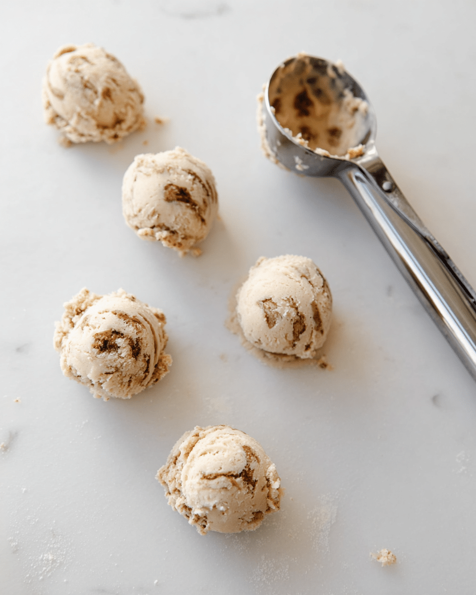 The image shows five small round scoops of light beige cookie dough with visible brown swirls and a slightly rough texture, placed evenly on a white marbled surface. A metal ice cream scoop with some dough stuck inside is positioned near the top right corner. The dough scoops look soft and slightly crumbly, spaced apart to avoid touching. photo taken with an iphone --ar 4:5 --v 7
