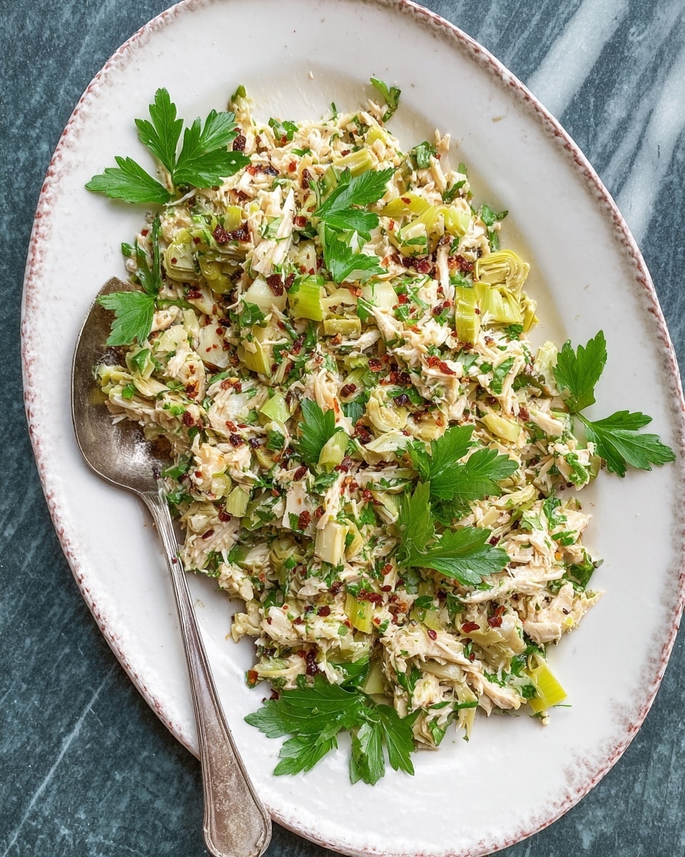 A close-up of a shredded chicken salad on a white plate with blue pattern edges, placed on a white marbled textured surface. The salad shows many layers of shredded pale beige chicken mixed with small pieces of light green celery and herbs. Bright green parsley leaves are scattered unevenly on top as garnish, adding fresh color. The texture looks moist with finely chopped small bits evenly distributed through the salad. A silver fork is placed near the edge of the plate on the right side. Photo taken with an iphone --ar 4:5 --v 7