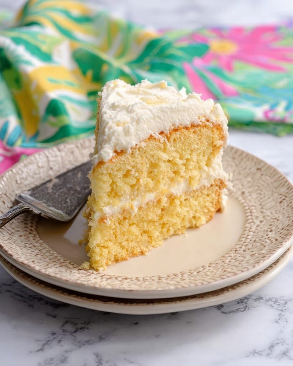 A large dome-shaped dessert with a rough, creamy yellowish outer layer sits on a white scalloped cake stand. On the top center of the dome, there is a small amount of light yellow garnish that looks like grated or chopped fruit. The stand is placed on a white marbled textured surface, and to the right side, there is a colorful cloth with pink, blue, yellow, and green patterns, next to an ornate silver serving spatula. The background is made of light gray stone tiles with a simple and clean look. photo taken with an iphone --ar 4:5 --v 7