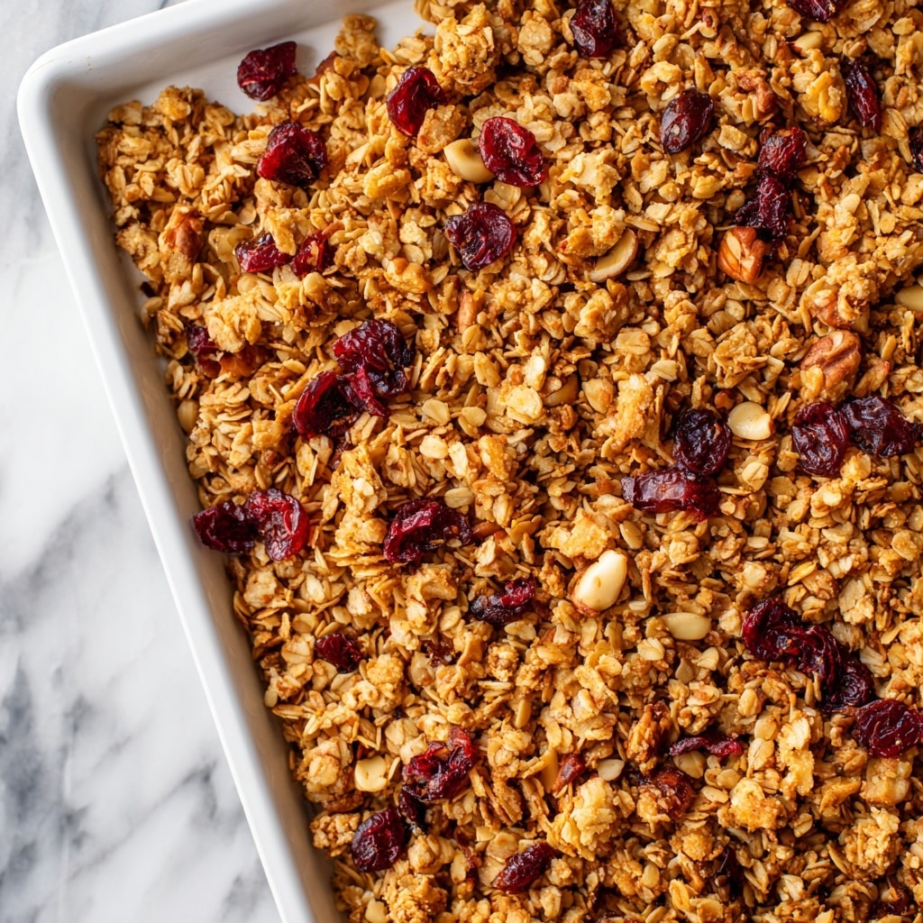 A close-up view of crunchy granola spread evenly across a white baking tray, showing a mix of golden brown oats, light and dark nut pieces, and clusters that give a rough texture. Scattered throughout are small, deep red dried berries adding contrast to the warm browns. The tray rests on a white marbled surface, and the granola appears toasted with a slightly shiny finish in parts. photo taken with an iphone --ar 4:5 --v 7