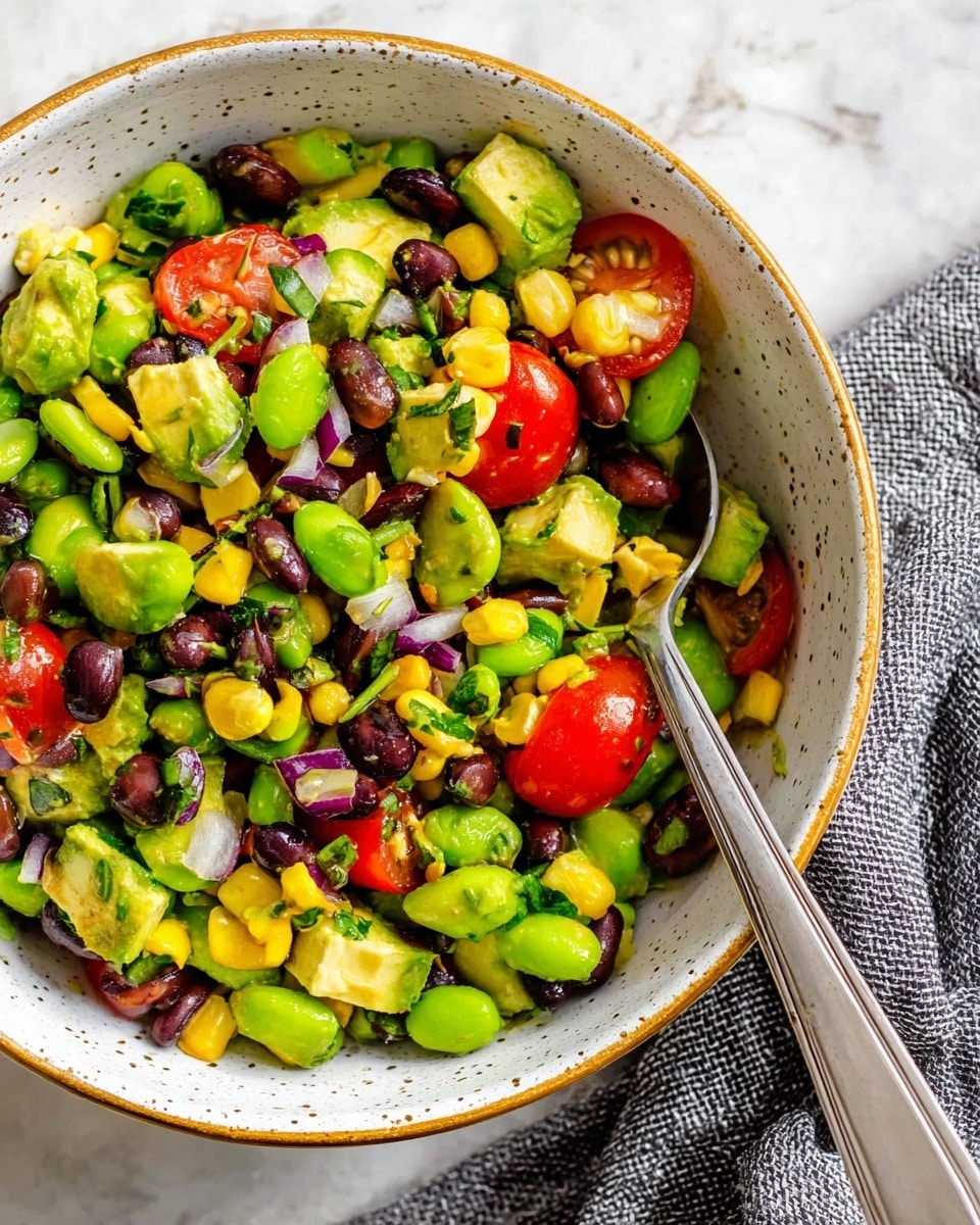A close-up view of a salad in a white speckled bowl with a thin golden rim, filled with four main layers: bright green chunks of avocado, light green edamame beans scattered throughout, shiny black beans, and yellow corn kernels. Red grape tomato halves add a pop of color, mixed with finely chopped purple onion and green herbs. A silver spoon rests inside the bowl on the right side, partially covered by the salad. The bowl is placed on a white marbled textured surface with a gray knitted cloth beside it. Photo taken with an iphone --ar 4:5 --v 7
