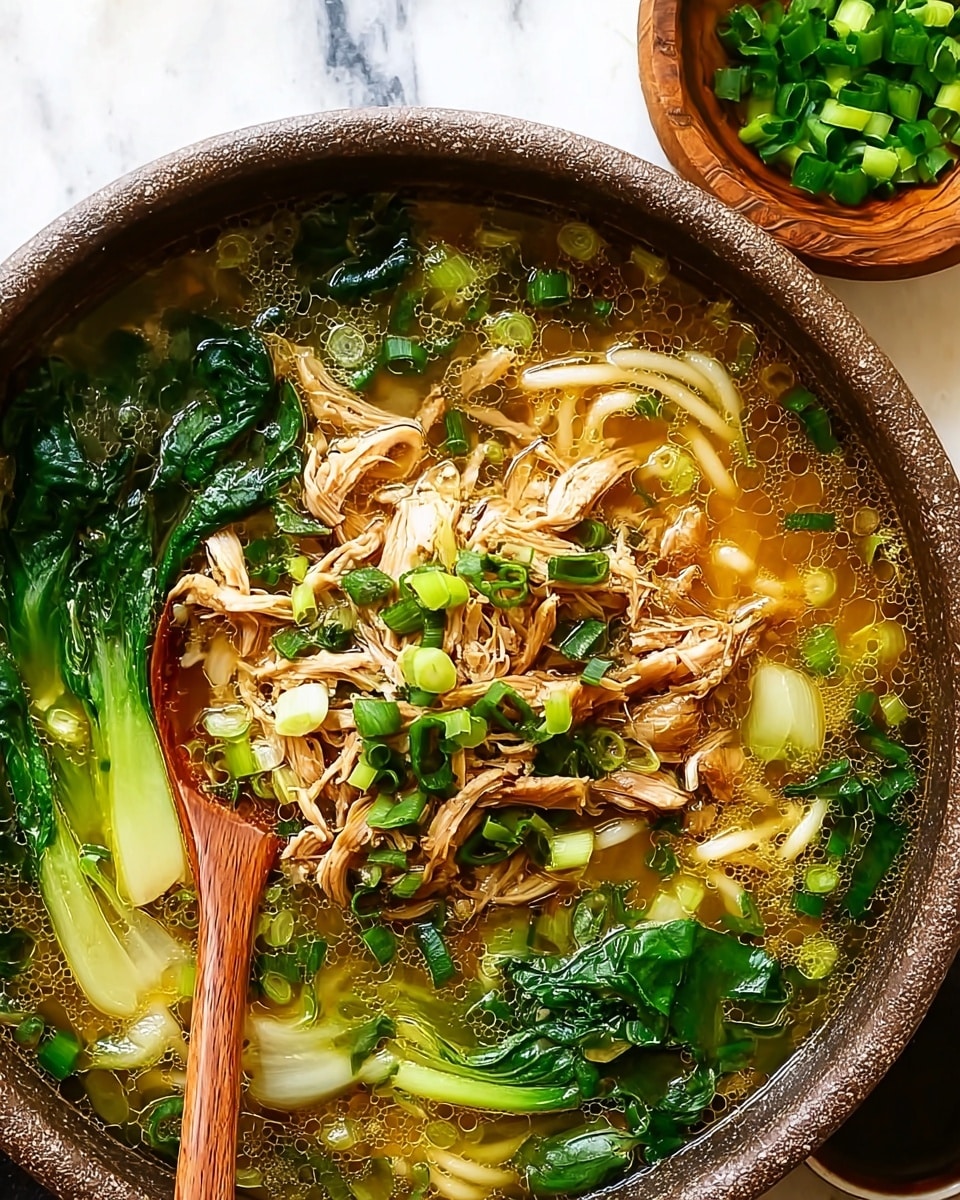 This image shows a bowl of soup with several layers. On the bottom, there is a clear golden broth with small oil bubbles. Inside the broth, there are thick white noodles twisted and partially submerged. On top of the noodles, there are green vegetables like bok choy with bright green leaves and pale green stalks. Scattered around are chopped green onions adding a fresh green touch. In the middle, there is a pile of shredded cooked chicken pieces that are light brown. A wooden spoon is partly dipped into the soup on the right side of the bowl. The bowl is white with a black speckled rim, placed on a white marbled surface. Photo taken with an iphone --ar 4:5 --v 7