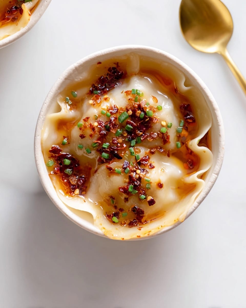 A close-up view of a single round dumpling in a white bowl, showing a smooth, slightly translucent dough with crinkled edges that fold over the filling. The dumpling is topped with glossy, reddish-brown chili oil pooling slightly around the edges and small bits of dark red chili flakes scattered across the surface. Bright green finely chopped chives are sprinkled on top, adding a fresh pop of color. The bowl sits on a white marbled surface, with a gold spoon placed to the right side. The overall look is clean and simple, highlighting the soft texture of the dumpling and the vibrant, spicy toppings. Photo taken with an iphone --ar 4:5 --v 7