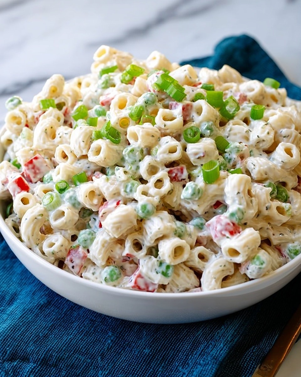 The image shows a large white bowl filled with a creamy pasta salad made of small, tube-shaped pasta mixed with a white dressing. The salad includes small pieces of green onions and finely chopped red and orange vegetables, adding bits of color throughout. The pasta looks soft and well-coated in the creamy sauce, and the green onion pieces give a fresh touch on top. The bowl sits on a white marbled surface with a dark blue cloth partly visible underneath. The photo is clear, bright, and focused closely on the salad. photo taken with an iphone --ar 4:5 --v 7