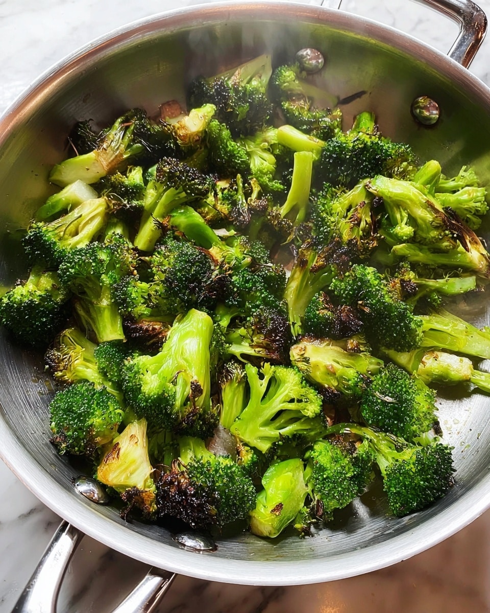 A close-up view inside a silver frying pan filled with cooked broccoli pieces that have a mix of bright green and slightly charred dark green colors, showing light browning on some edges. The broccoli pieces vary in size with visible florets and stems, and droplets of steam rise gently from the pan indicating heat. The pan has a reflective surface and rivets attaching the handle are visible. The scene is set on a white marbled textured surface. photo taken with an iphone --ar 4:5 --v 7