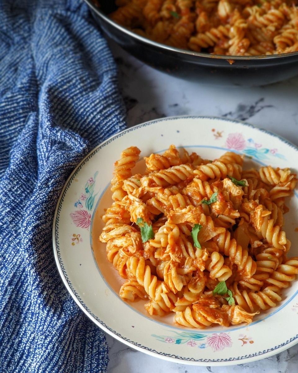 A white plate with delicate blue and pink floral patterns holds a serving of rotini pasta coated in a light red tomato sauce, mixed with shredded chicken pieces and garnished with small green cilantro leaves. The pasta appears glossy and slightly saucy, with the spirals of the rotini clearly defined. Behind the plate, a black pot filled with more of the same pasta dish is visible, and to the left, a textured blue fabric with white stripes is casually placed on a white marbled surface. photo taken with an iphone --ar 4:5 --v 7