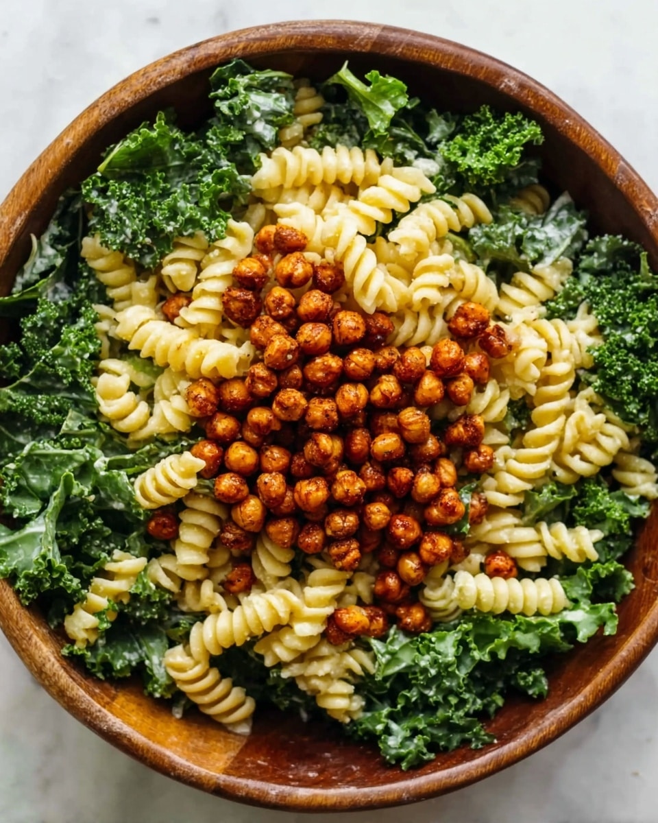 A wooden bowl filled with a salad showing three main layers: the bottom layer has curly green kale, the middle layer is light yellow rotini pasta, and the top layer is small reddish-brown roasted chickpeas scattered evenly. The textures include leafy and rough kale, smooth and spiral-shaped pasta, and crispy chickpeas. The photo is taken from above, showing the mix clearly on a white marbled surface. photo taken with an iphone --ar 4:5 --v 7