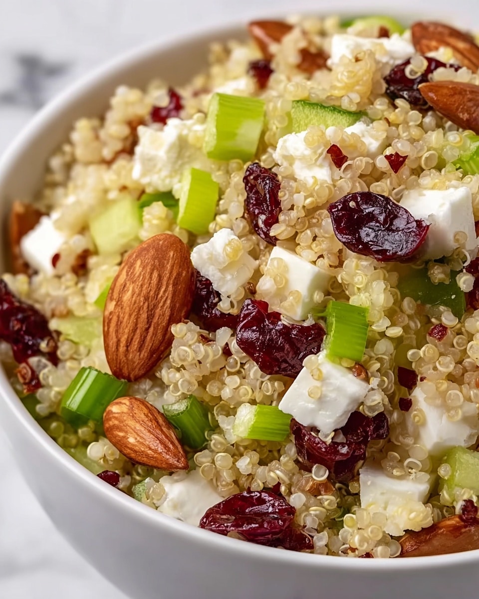 This image shows a close-up of a quinoa salad in a white bowl. The salad has several layers, starting with a base layer of light beige quinoa grains with a tiny red specks mixed in. Scattered on top are chunks of white feta cheese with a crumbly texture, pieces of green celery and green onions providing a fresh, crisp look, dried dark red cranberries adding a chewy texture, and whole and sliced brown and beige almonds for crunch. The ingredients are mixed evenly, creating a colorful and textured dish. The background is a white marbled texture. Photo taken with an iphone --ar 4:5 --v 7