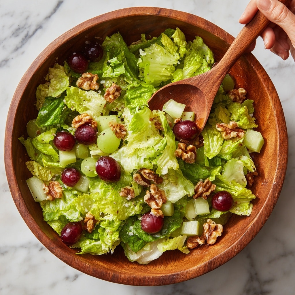 A wooden bowl filled with a fresh salad sits on a white marbled surface. The salad has three main layers: the bottom layer is light green leafy lettuce that looks soft and curly, the middle layer is thinly sliced pale green celery with a crunchy texture, and the top layer features bright green apple slices, small dark red grapes, and scattered light brown walnut pieces. Tiny bits of red dried fruit are sprinkled throughout, adding small pops of color. The salad looks fresh and crisp, with a mix of smooth, crunchy, and soft textures. photo taken with an iphone --ar 4:5 --v 7