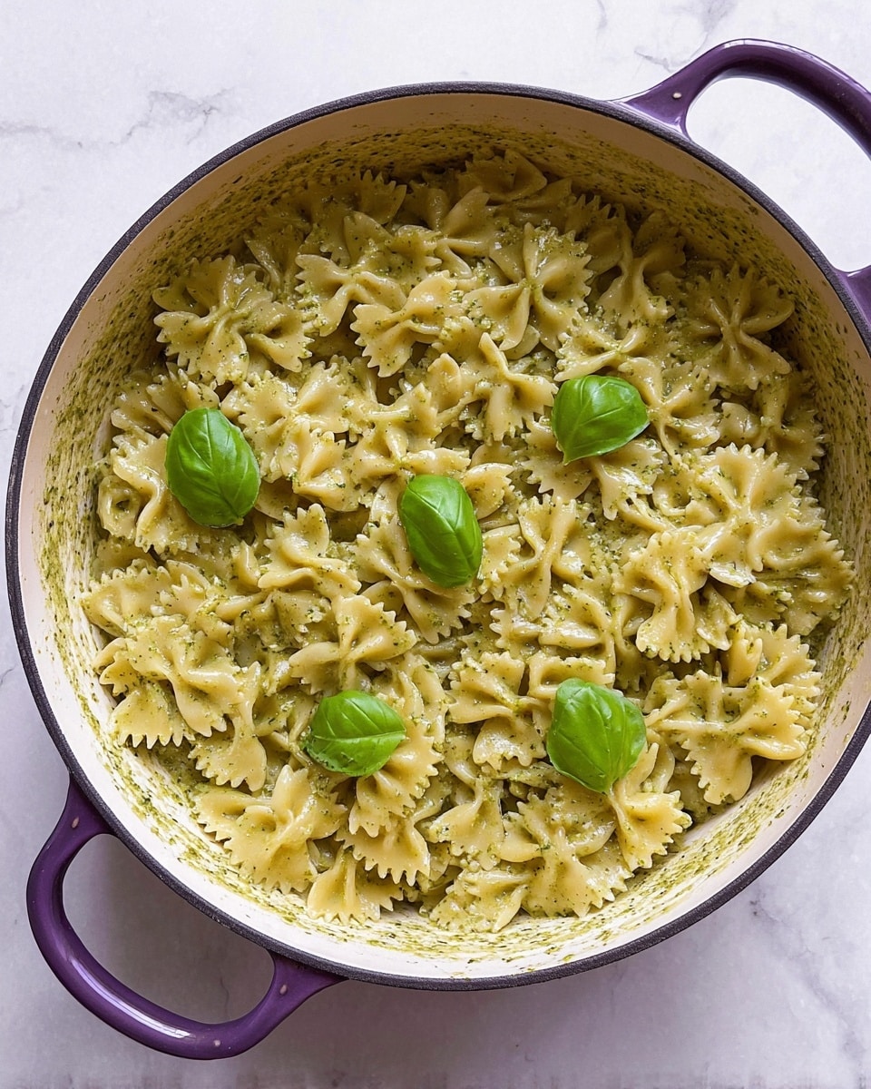A large white pot with purple handles is filled with a creamy pasta dish made of farfalle noodles coated evenly in a pale green pesto sauce with specks of herbs. The sauce gives the pasta a smooth, slightly glossy texture. On top of the pasta, several fresh green basil leaves are placed as garnish, adding a bright contrast to the creamy sauce and the light yellow noodles. The pot sits on a white marbled texture surface. photo taken with an iphone --ar 4:5 --v 7
