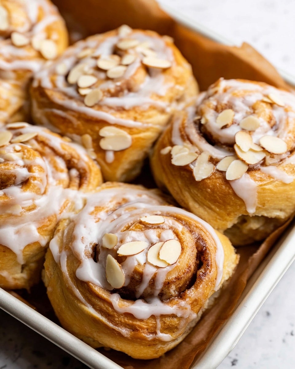 The image shows close-up light brown cinnamon rolls arranged in a baking dish, each roll featuring a visible spiral pattern with darker cinnamon filling inside. The rolls are topped with a thick white cream glaze that is gently drizzled over, slightly pooling around them. Scattered thin, off-white almond slices cover the tops, adding texture. The background is a white marbled surface with soft natural light highlighting the warm and creamy colors of the rolls and toppings. Photo taken with an iphone --ar 4:5 --v 7