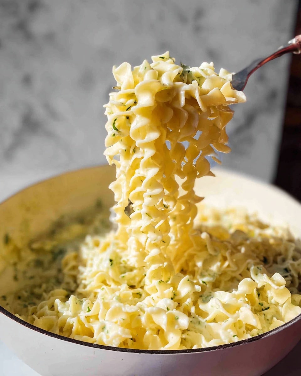 The image shows a close-up of a pan filled with ruffled, ribbon-like pasta coated in a light green, creamy sauce with small herb pieces, giving the sauce a speckled texture. The pasta strands are curly, pale yellow, and slightly overlapped, showing the smooth, saucy surface mixed with bits of green herbs evenly spread throughout. The inside of the pan is light cream, and the scene is set against a white marbled texture. photo taken with an iphone --ar 4:5 --v 7