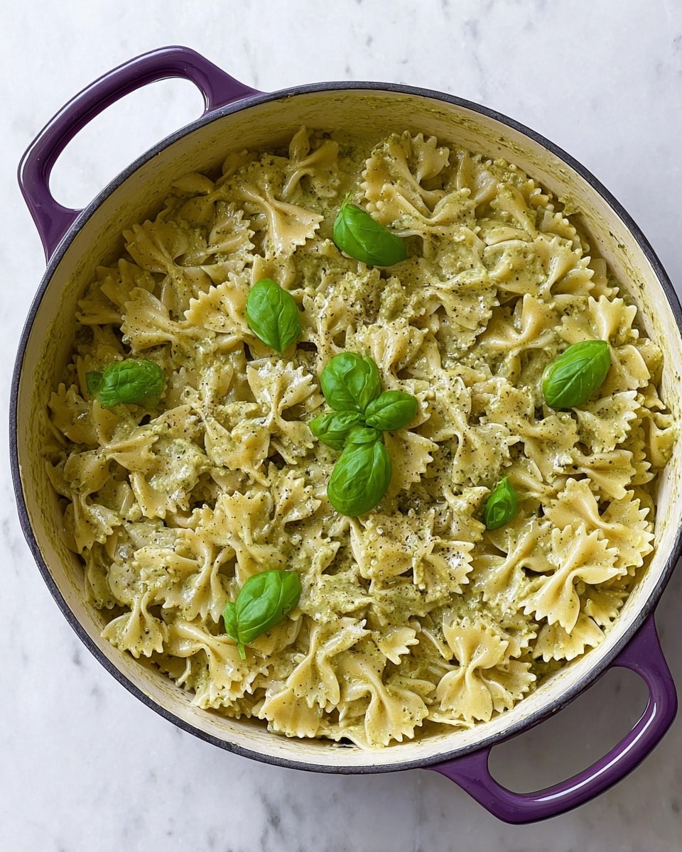 A large round purple pot filled with one layer of creamy pesto farfalle pasta, the light green sauce coating each pasta piece evenly with small flecks of herbs. On top, four bright green basil leaves are placed in small clusters. The pot sits on a white marbled surface showing some sauce remnants along the pot's inner edge. photo taken with an iphone --ar 4:5 --v 7